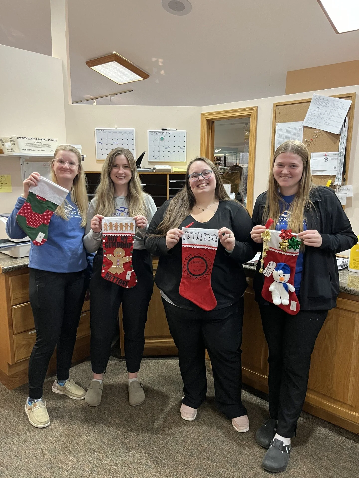 Four women standing behind a counter, holding Christmas stockings with holiday-themed decorations, in an indoor office or break room with bulletin boards and a door in the background.