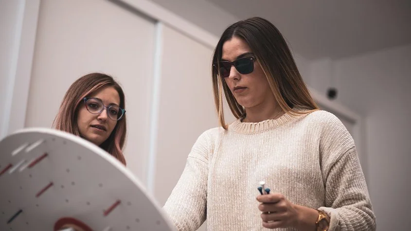 Two women with glasses, one with dark sunglasses, stand in a room looking at a large round object doing Vision therapy exercises.