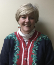 Smiling woman with short blonde hair wearing a festive Christmas sweater with holly design, standing against a plain wall. She is a Vision Therapy graduate from Superior Eye Health.