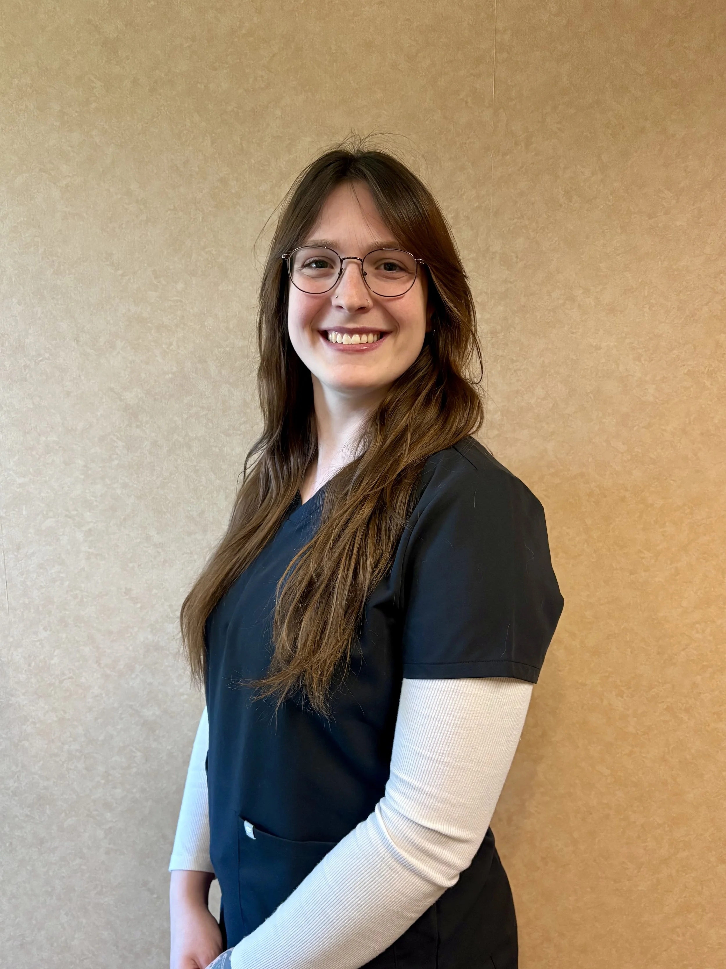 A woman with long brown hair, wearing glasses, a black top, and a beige long-sleeve shirt underneath, is smiling and standing against a plain beige wall.