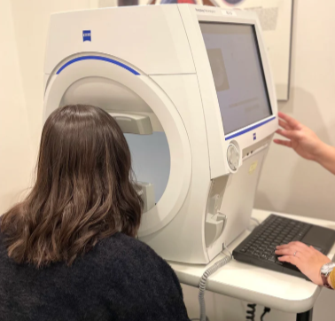 A woman undergoing an eye exam using a large, modern automated visual field testing machine in a clinical setting, with a technician operating the machine.