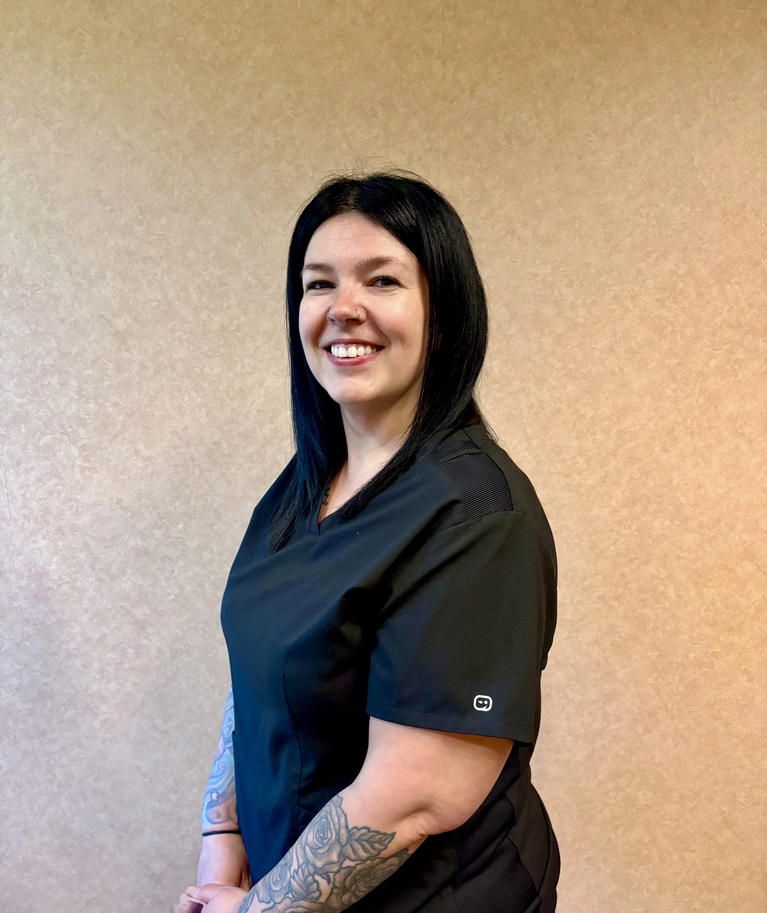 A woman with black hair and a nose piercing, wearing a black shirt, stands against a beige wall, smiling at the camera with her arms crossed, showcasing a tattoo on her left forearm.