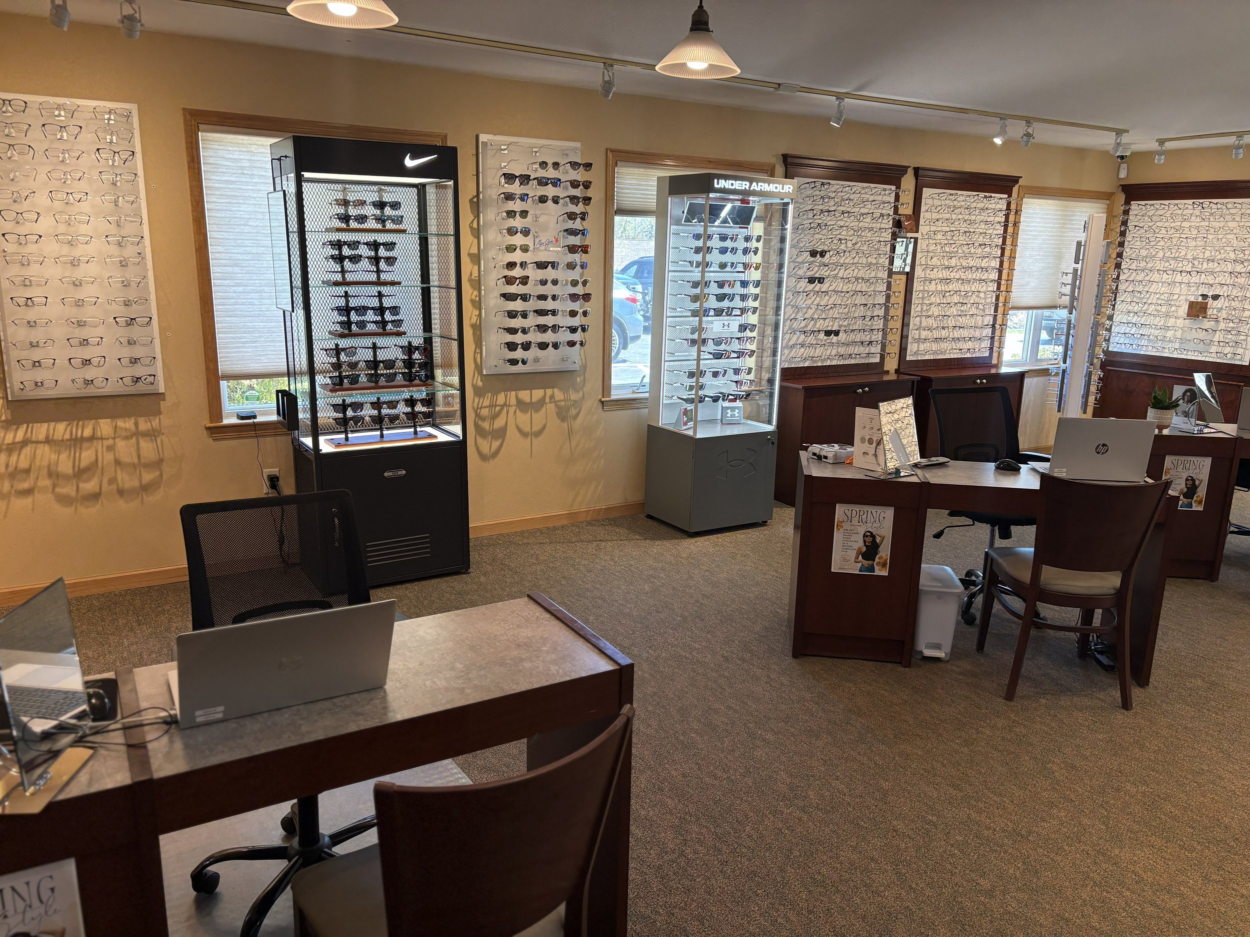 Interior of an eyewear store with display walls of glasses, sunglasses, and contact lenses, a desk with a laptop, and chairs, with windows letting in natural light.