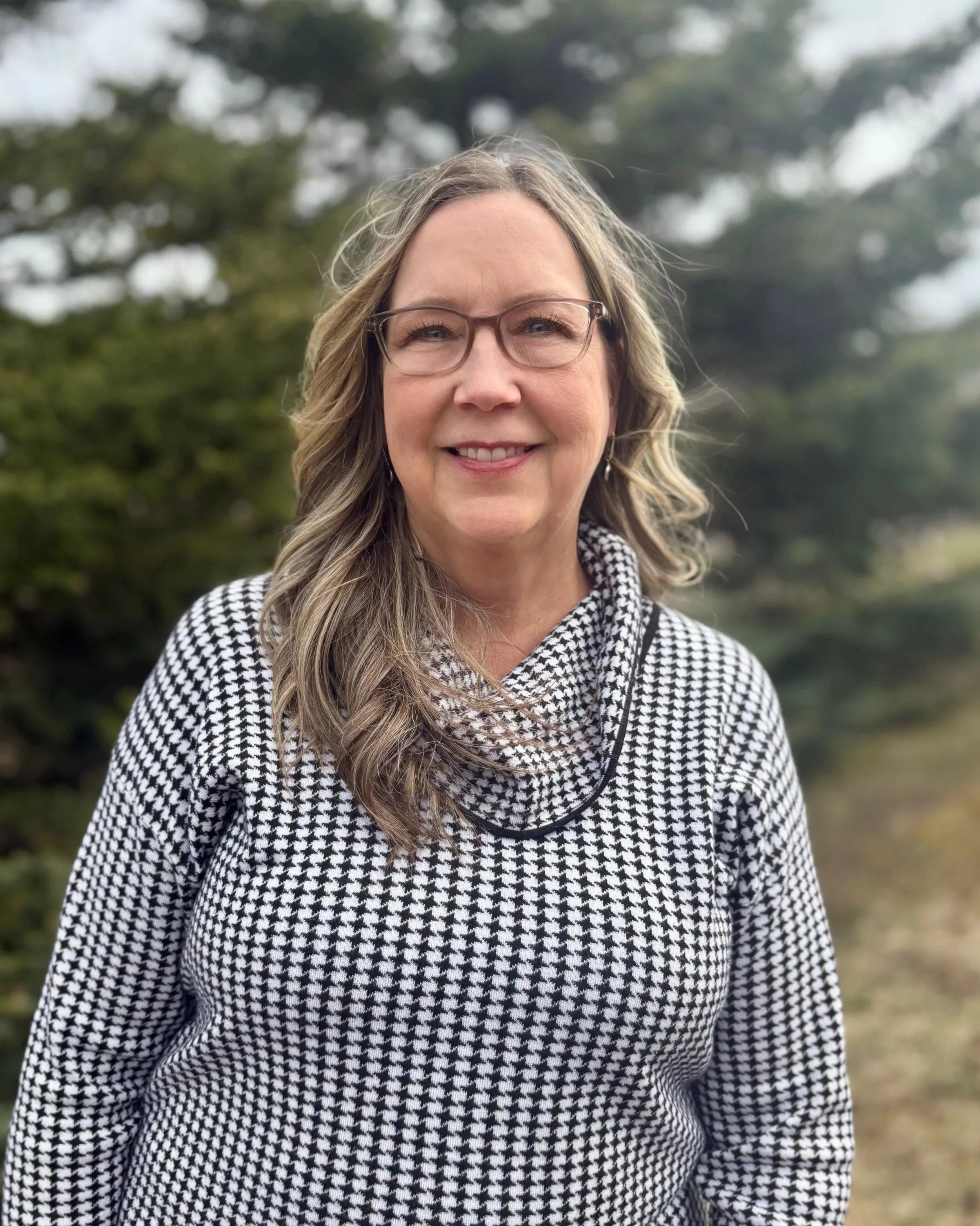 A woman with long wavy blonde hair, glasses, and earrings smiling outdoors in front of a blurred background of trees, wearing a black-and-white houndstooth patterned coat.