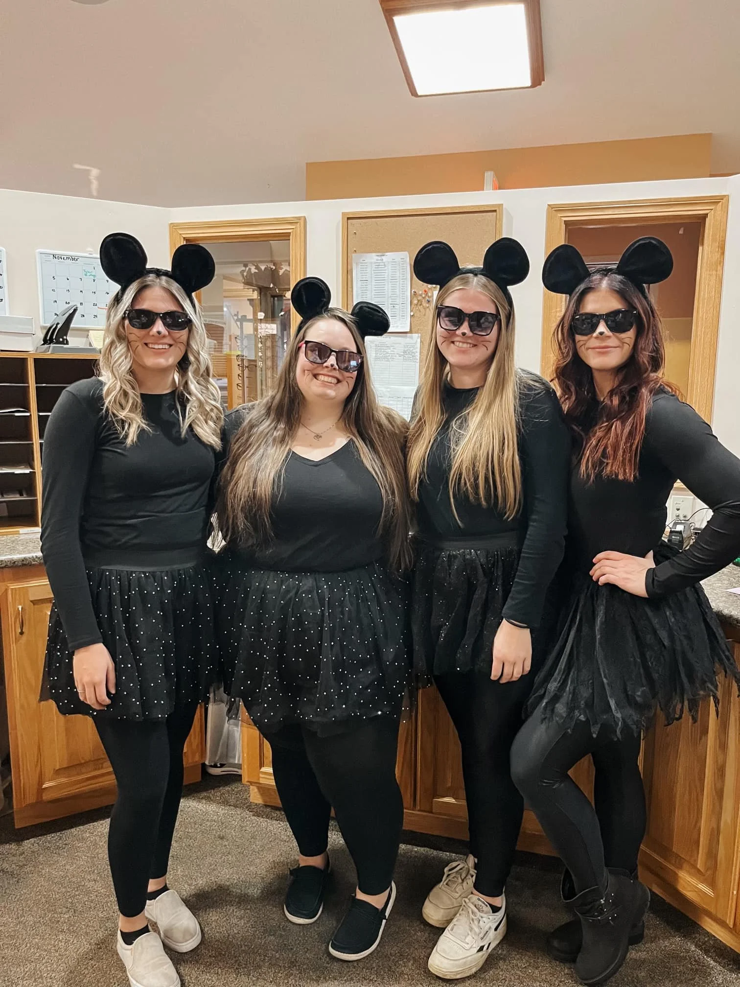 Four women dressed in black with black mouse ears headbands, sunglasses, and black skirts with white polka dots, posing together in a room with wooden cabinetry, a whiteboard, and some papers on the wall.
