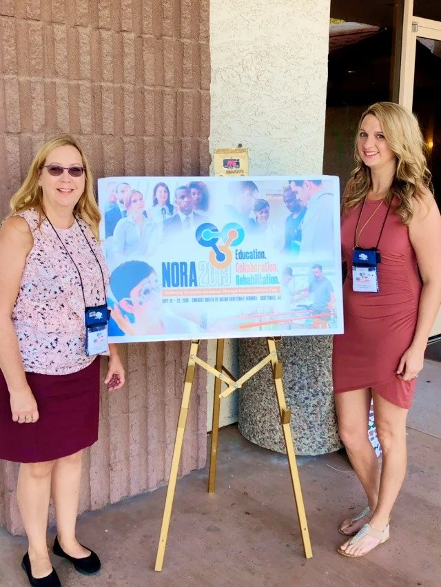 Two women standing next to a promotional sign on an easel at a conference. The sign has the text 'NORA 20' and mentions education, collaboration, and rehabilitation.