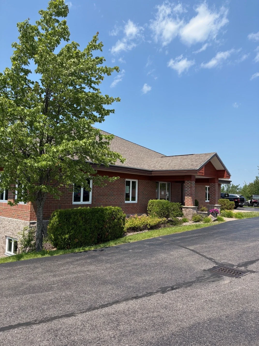 A brick building in Marquette, Michigan with a gabled roof, surrounded by green bushes and trees, with a parking lot and a clear blue sky in the background.