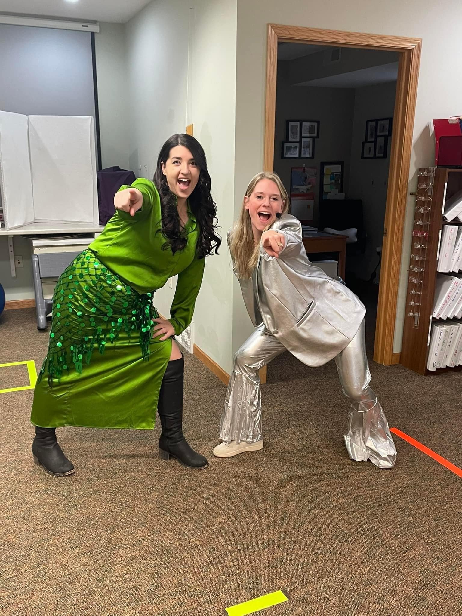Two women dressed in colorful, shiny costumes posing playfully in an indoor office setting.