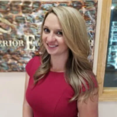 A woman, Dr. Jessica Jackson, with blonde hair wearing a pink top, smiling, in front of a map or textured wall at Superior Eye Health Center.