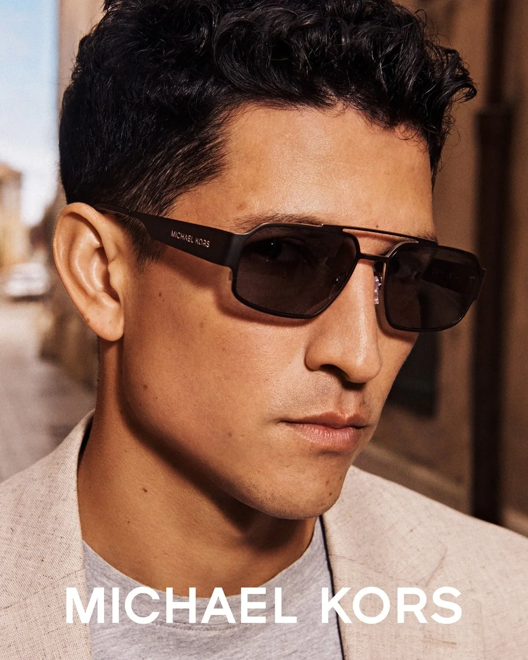 Close-up of a man with short curly hair wearing black Michael Kors sunglasses and a light-colored blazer, with the brand name 'MICHAEL KORS' visible on the sunglasses arm and at the bottom of the image.