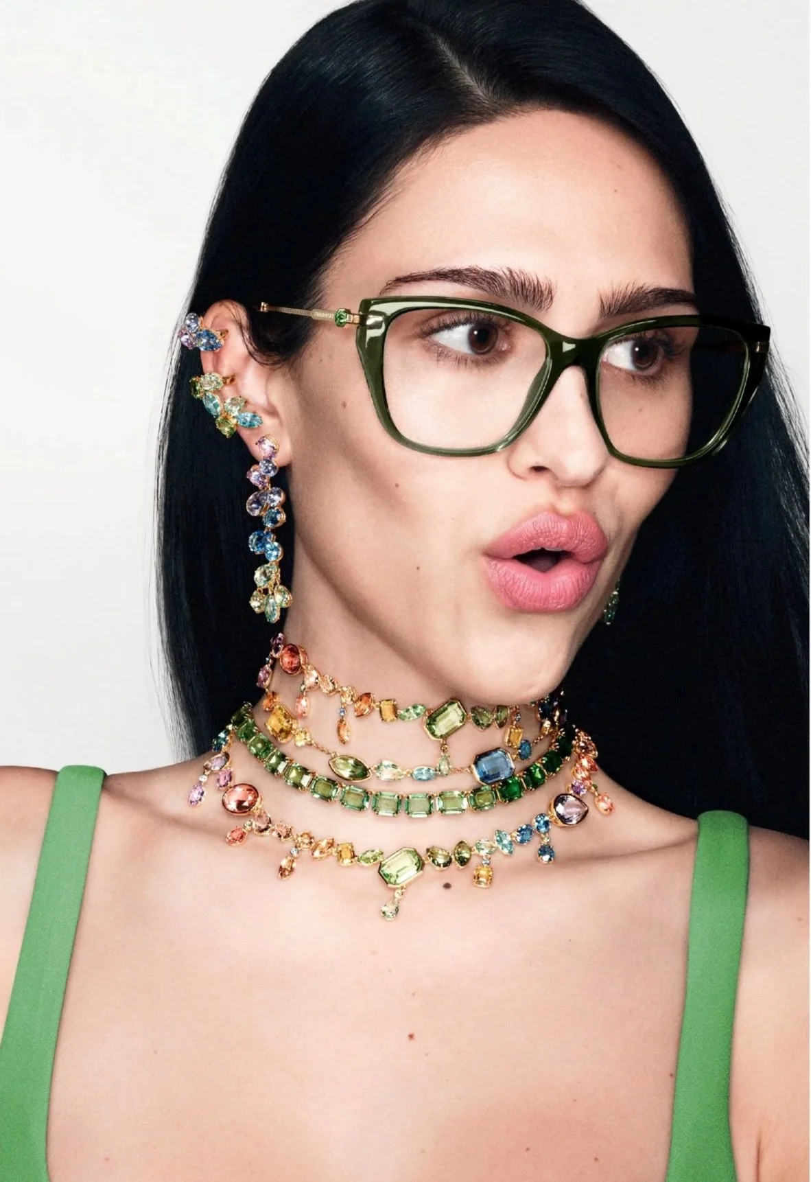 A woman with black hair wearing Swarovski green glasses, colorful earrings, and multiple layered colorful gemstone necklaces, with a green strap top.