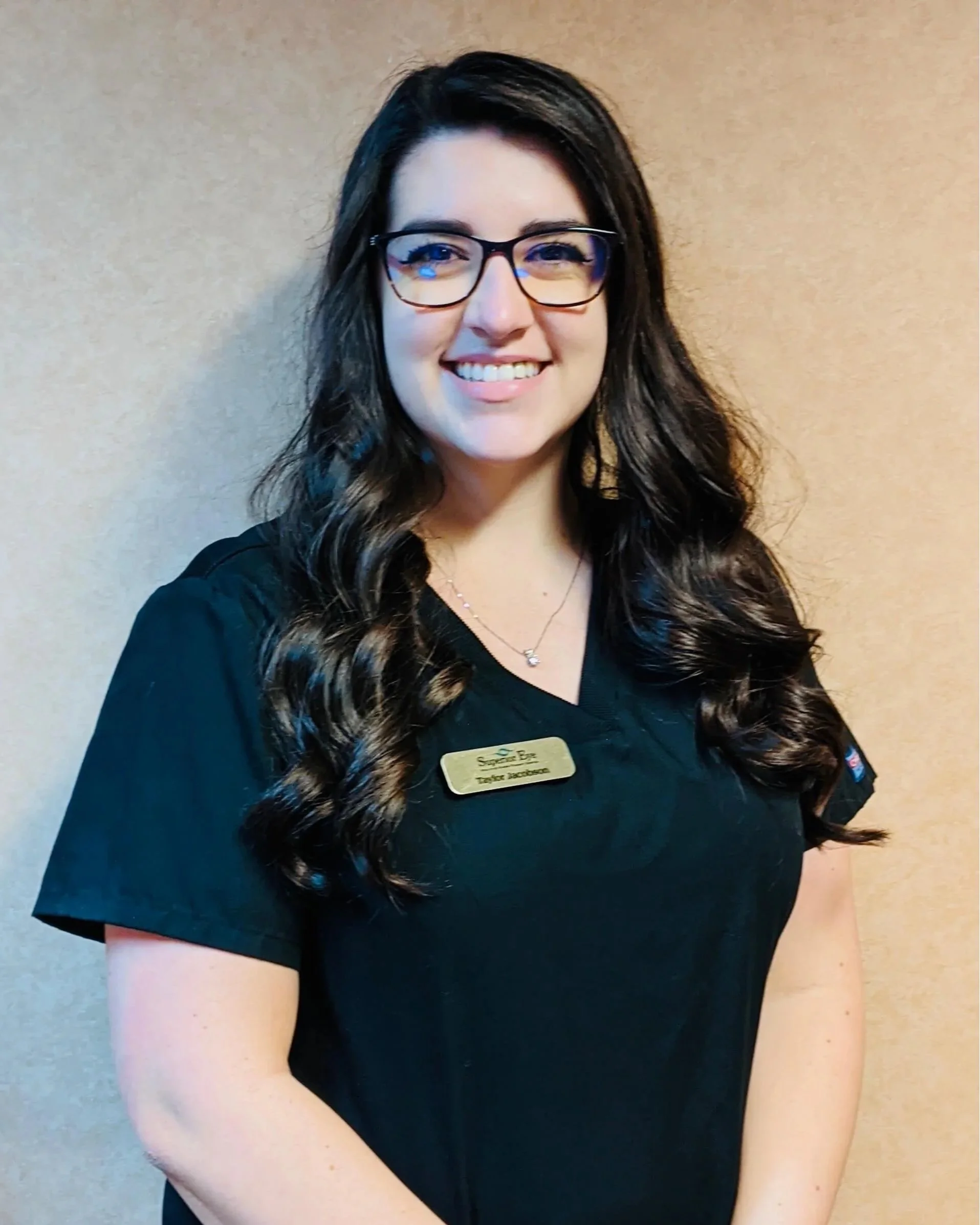 Close-up of a smiling woman with dark brown hair, glasses, and a name tag, standing against a neutral beige background.
