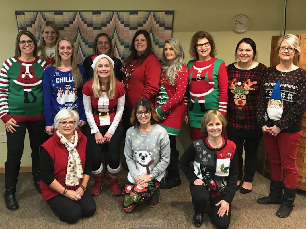 Group of people wearing Christmas sweaters, some with holiday decorations, smiling in a room with a quilted wall hanging and a clock.