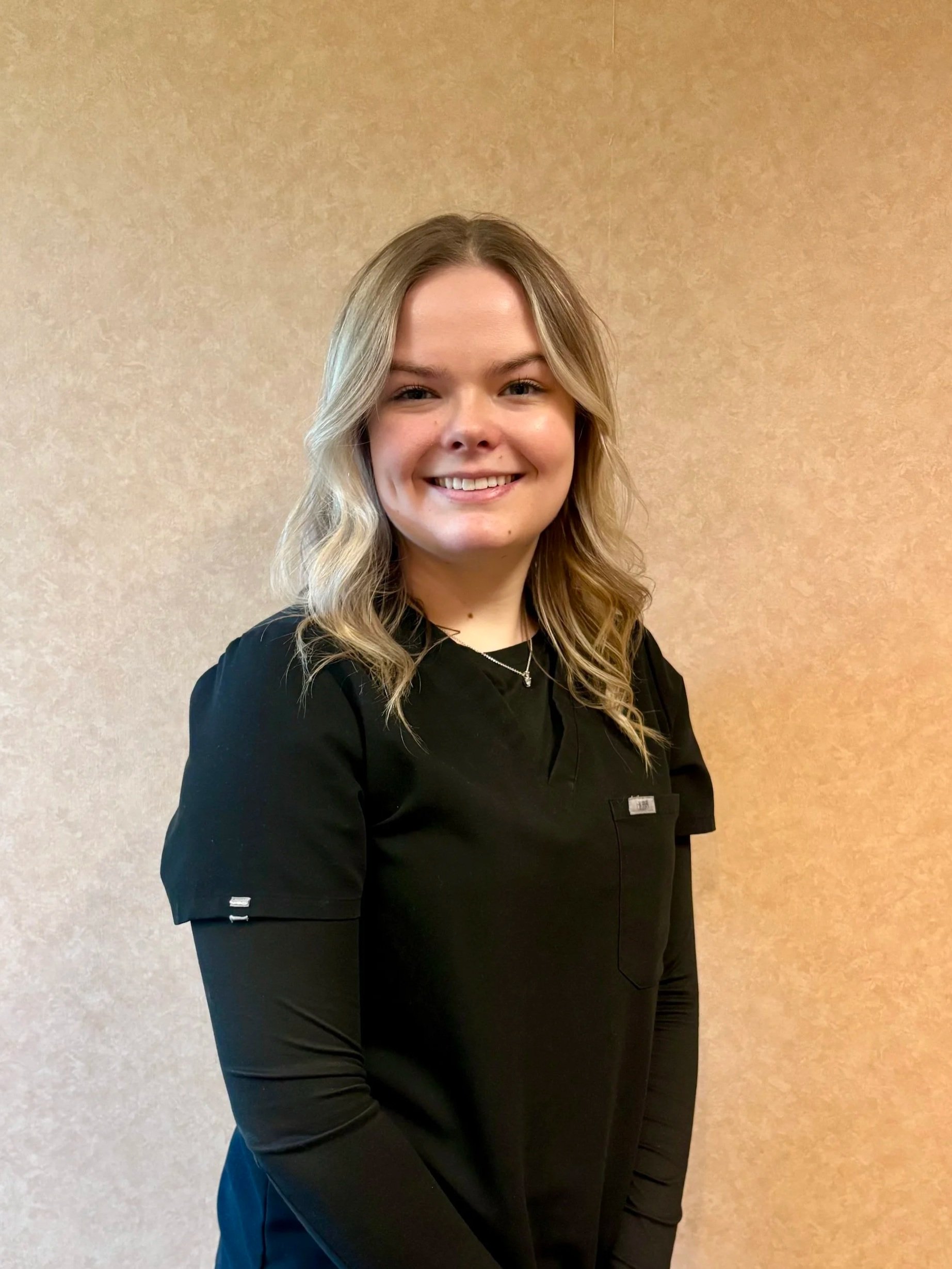 A young woman with long, wavy blonde hair smiling, wearing a black uniform, standing in front of a beige wall.