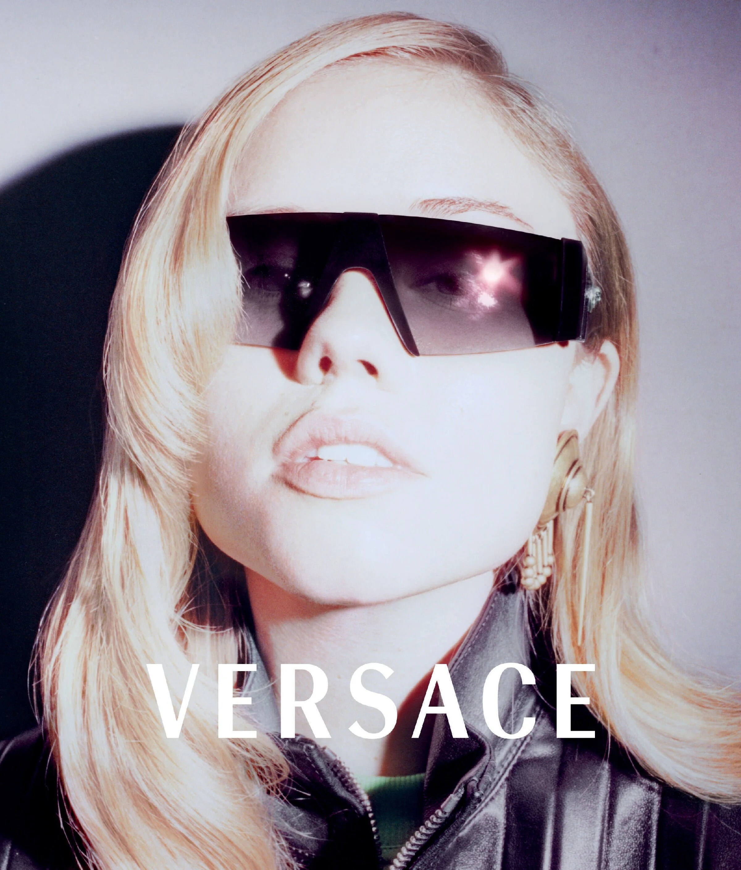 Close-up of a woman wearing large black sunglasses with reflective lenses, blonde hair, and a black leather jacket, with the word "VERSACE" overlaid at the bottom.