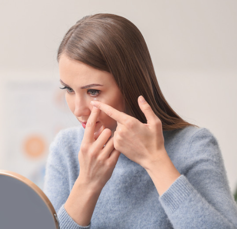 Woman touching her eye while looking into a mirror, putting in a contact lens.