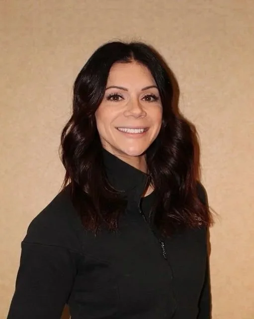 A woman with dark wavy hair smiling, wearing a black zip-up jacket, standing against a beige wall.