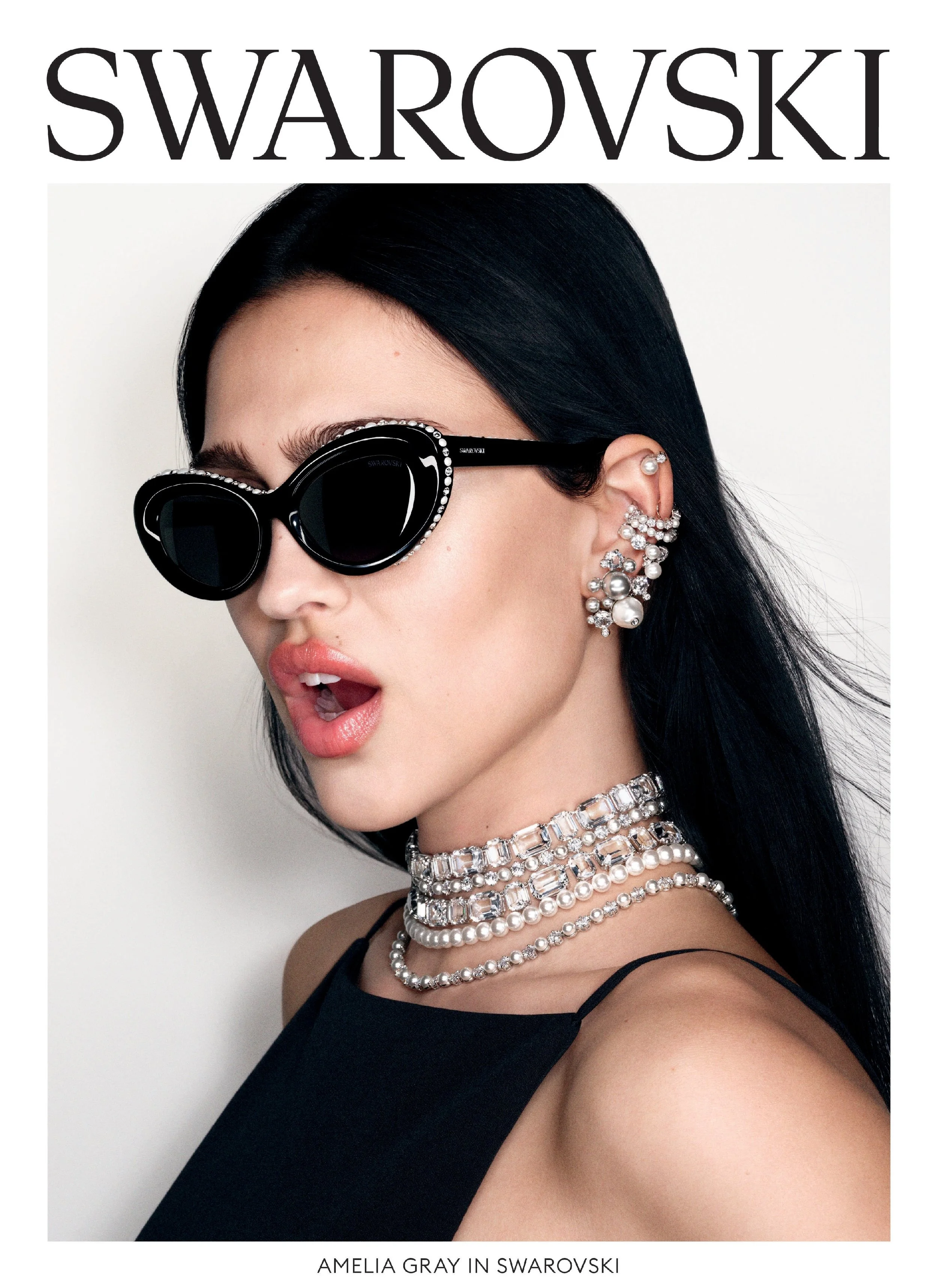 A woman wearing black sunglasses with Swarovski embellishments, multiple pearl and crystal necklaces, and matching earrings. She has black hair and is dressed in black, posing against a white background.