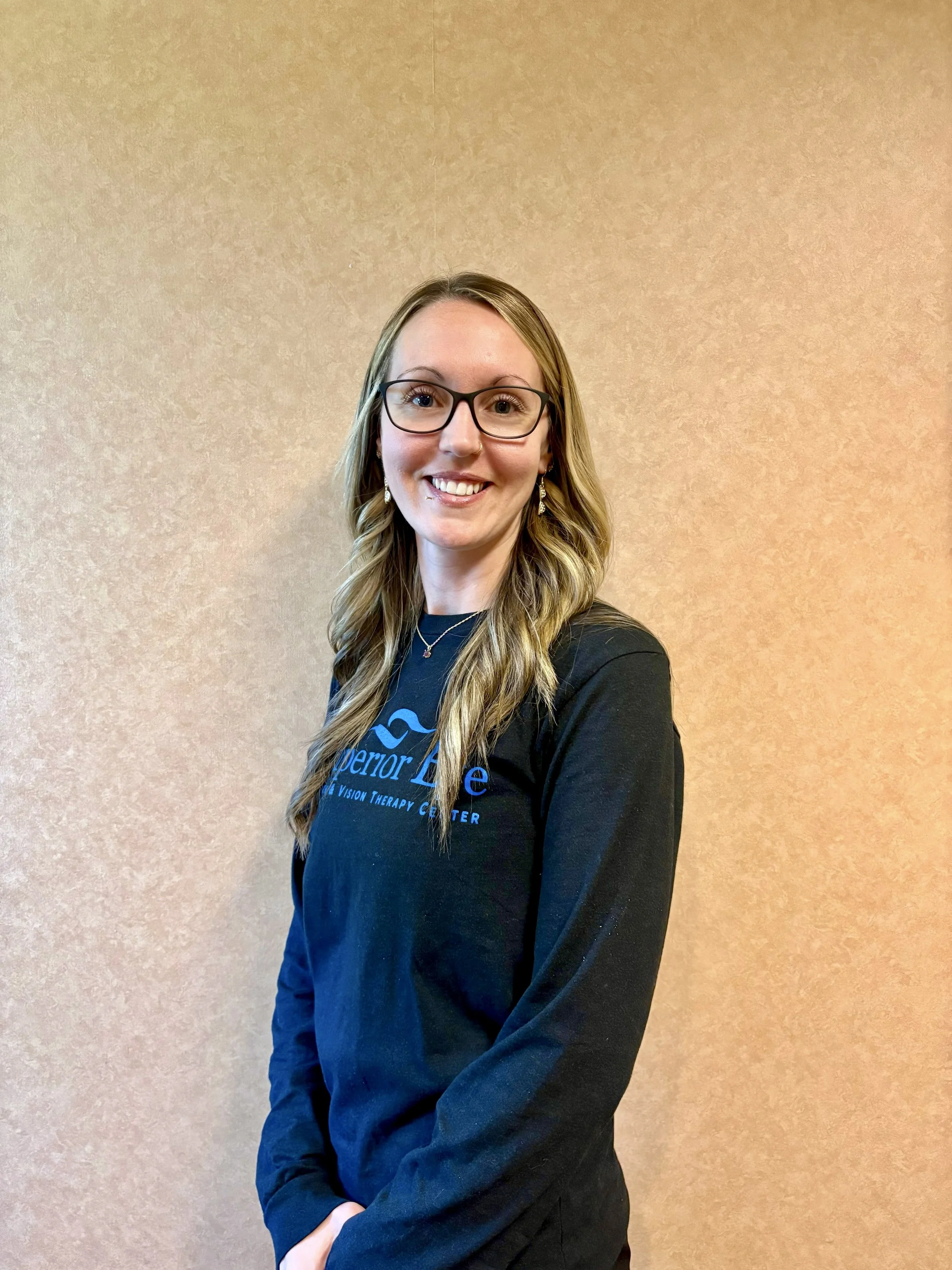 A young woman with long, wavy hair wearing glasses, earrings, and a black shirt with a logo that says 'Superior Eye' stands in front of a plain beige wall. She is smiling and looking directly at the camera.