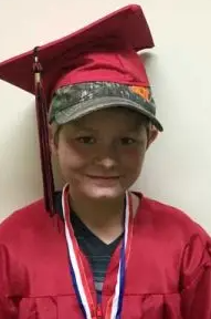 Young child in a red graduation gown and cap, wearing a camouflage hat underneath, smiling at the camera. He is a Vision Therapy graduate from Superior Eye Health.