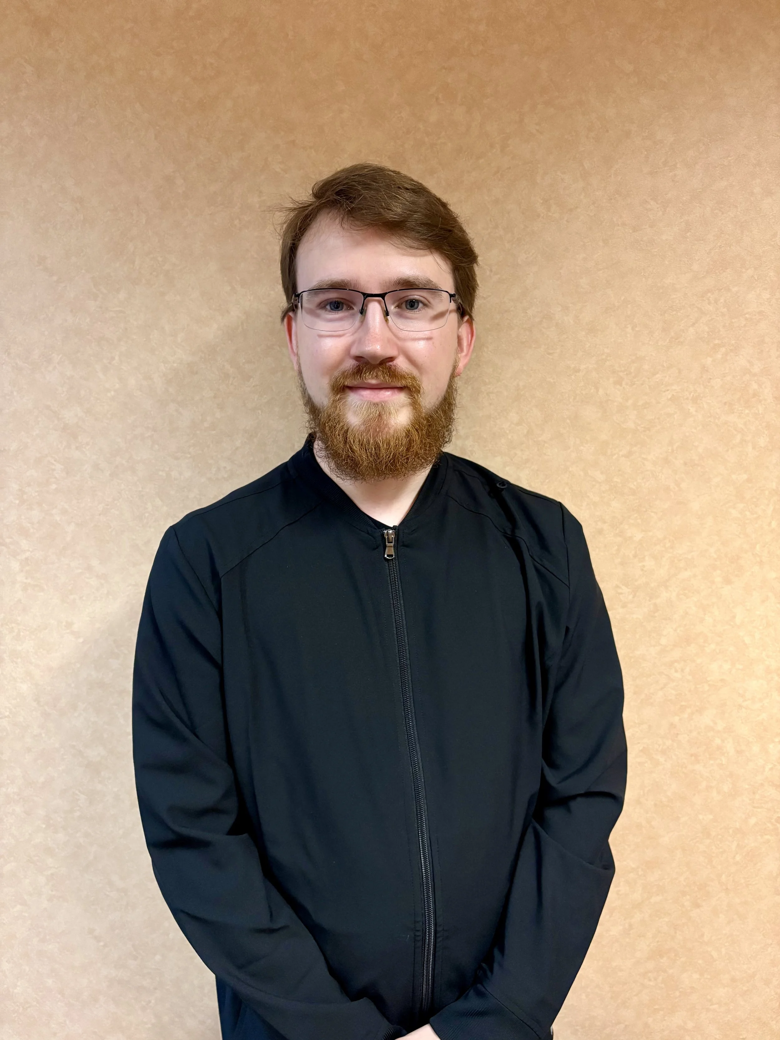 A man with glasses, a beard, and brown hair, wearing a black zip-up jacket, stands against a beige textured wall, smiling softly at the camera.