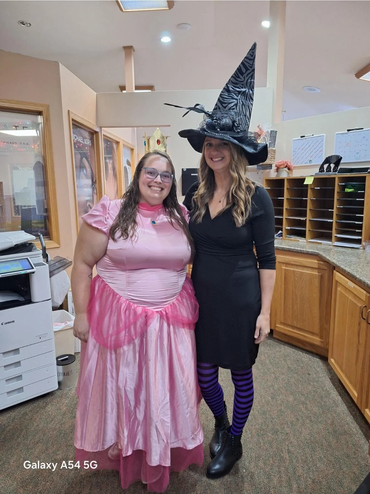 Two women smiling indoors, one dressed as a princess in a pink gown and crown, the other in a black dress with a large witch hat and purple striped tights.