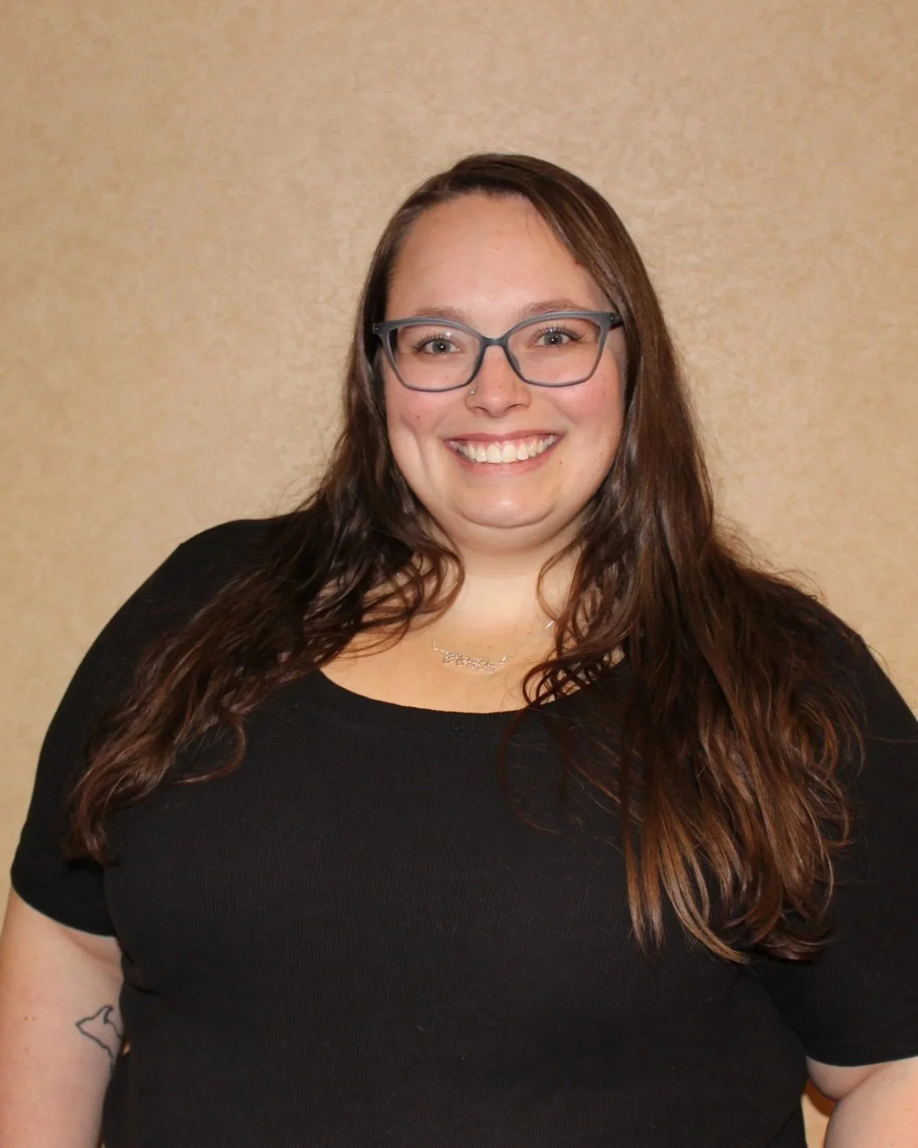 A woman with long wavy brown hair, wearing glasses, smiling, standing against a beige background, dressed in a black top, with a small tattoo on her left arm and a necklace
