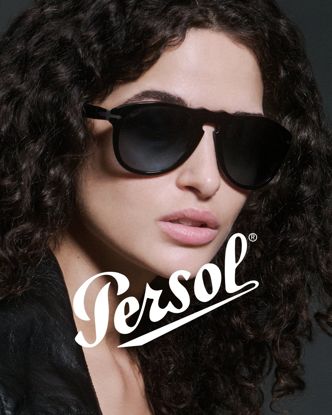 A woman with curly dark hair wearing large black sunglasses and a black leather jacket, with the Persol logo overlayed on the image.