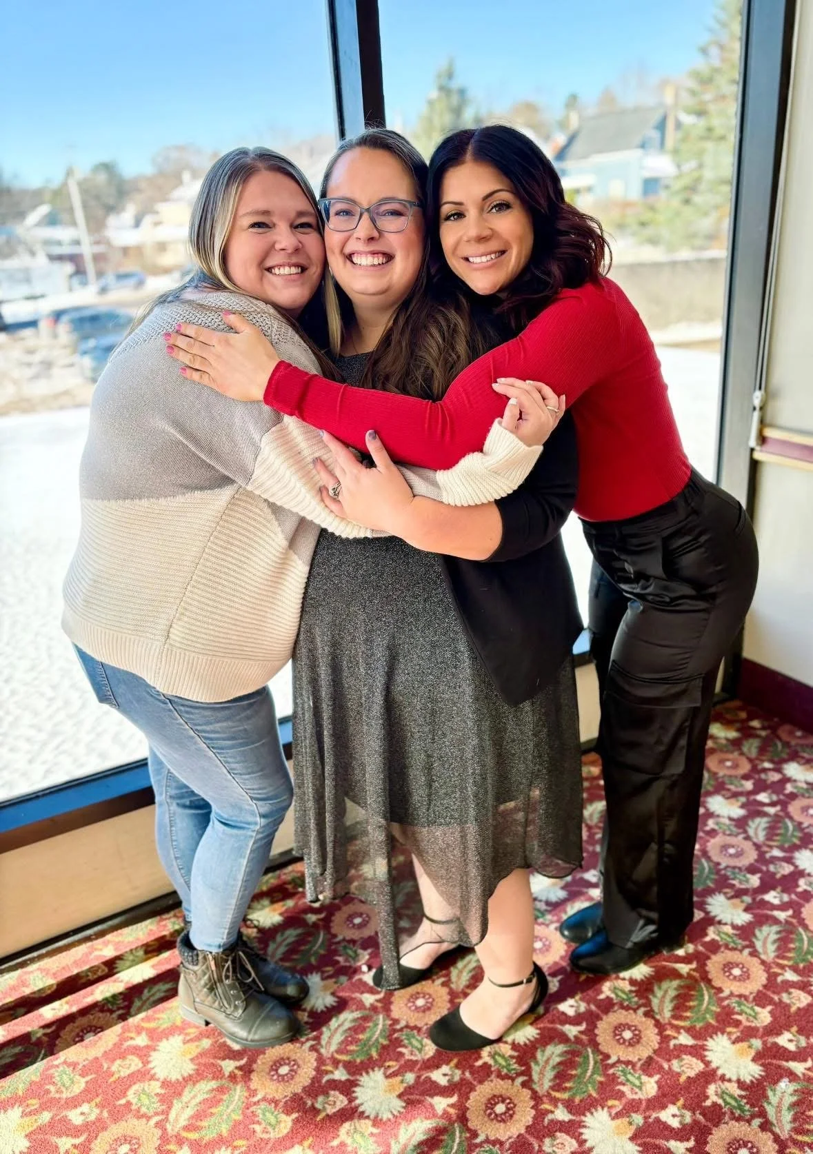 Three women hugging indoors near a large window with a view of houses and trees in the background. They are smiling and wearing casual and semi-formal clothing, including sweaters, a dress, and shiny pants.