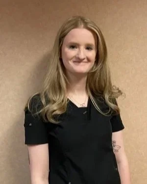 A young woman with long, wavy blonde hair, wearing a black shirt, standing against a beige background and smiling.