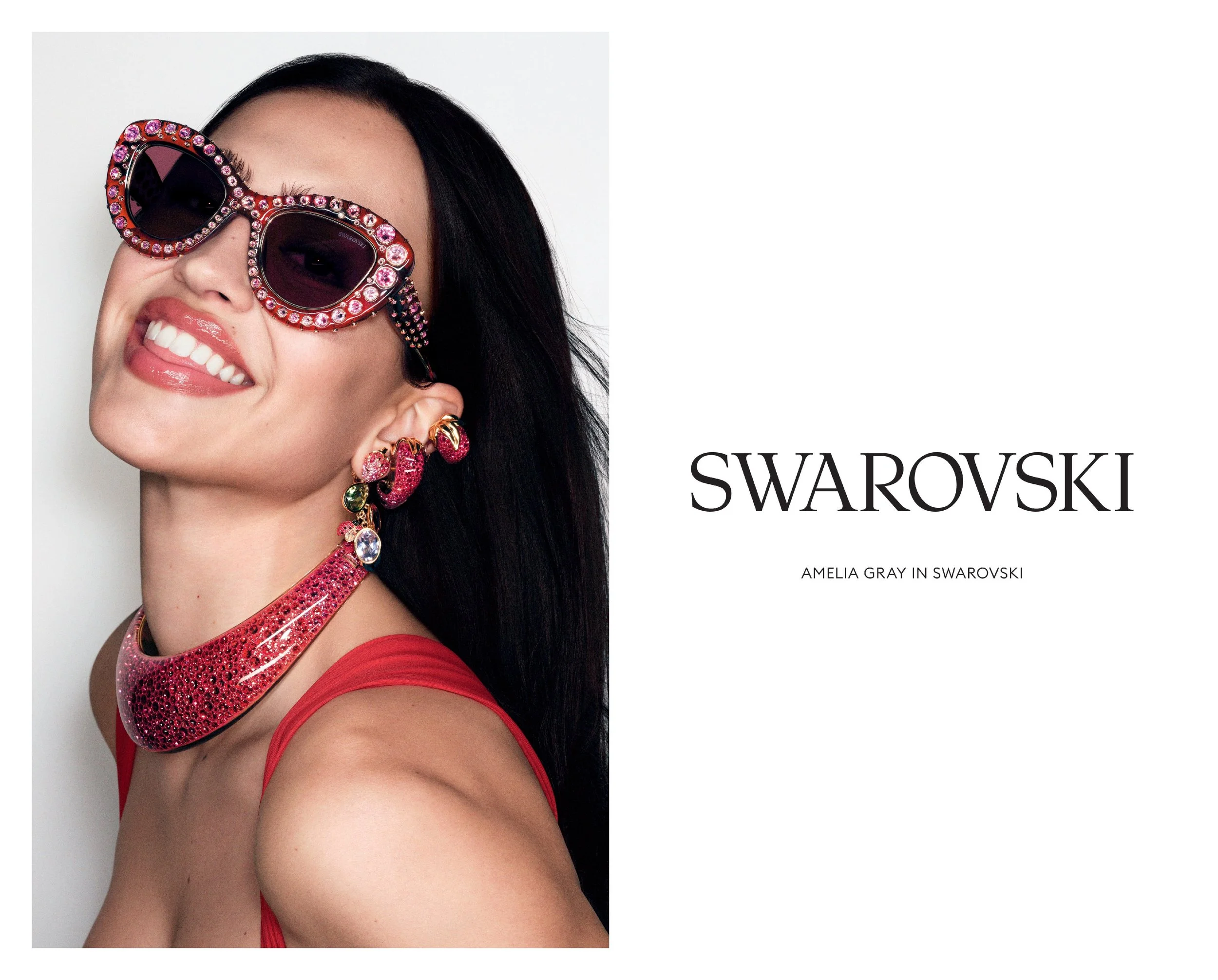 A woman wearing large, pink, and black sunglasses with rhinestones, red lipstick, and colorful jewelry, smiling against a plain background.