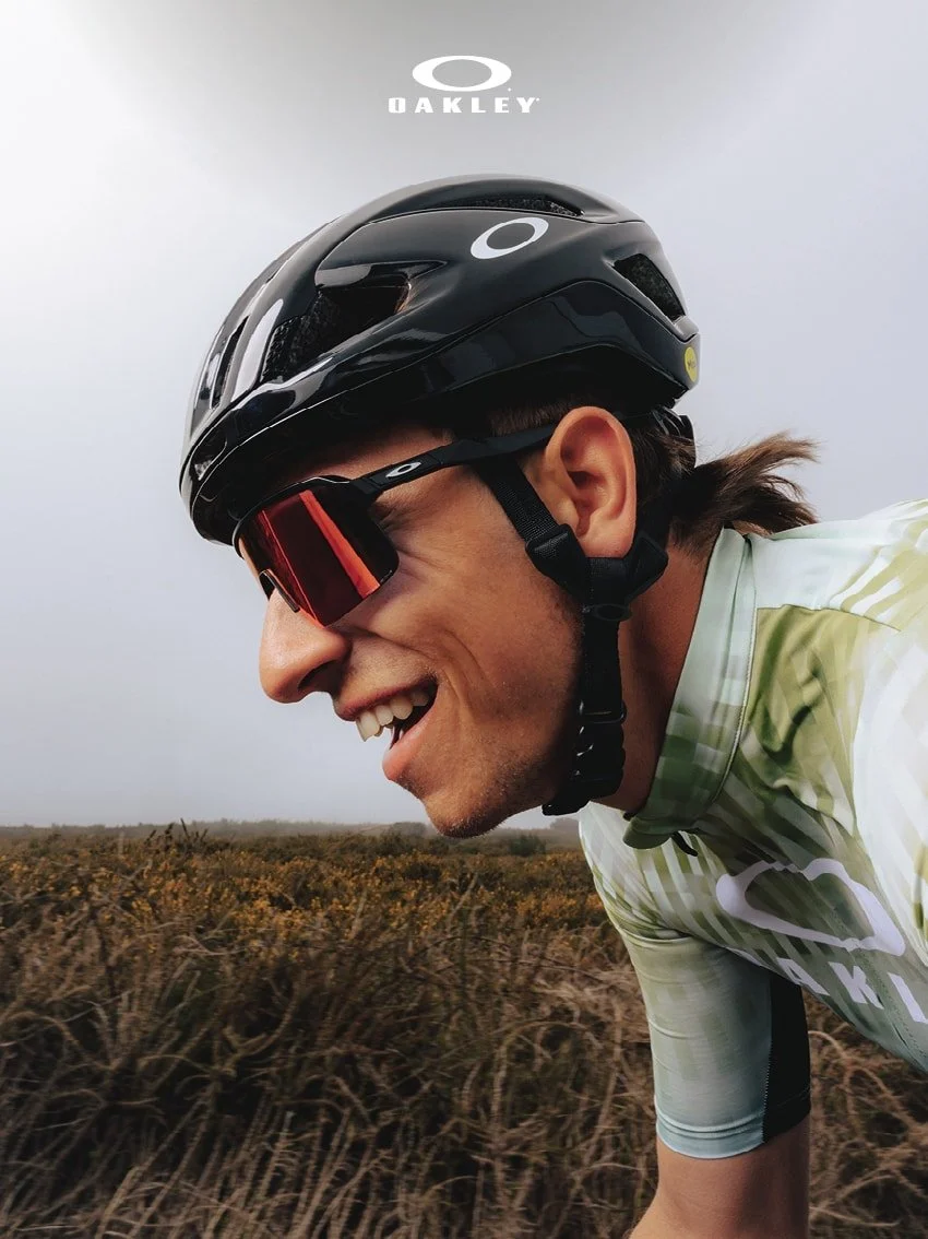 Close-up of a cyclist wearing a black helmet and red sunglasses, smiling while riding through a field of tall grass on a cloudy day.