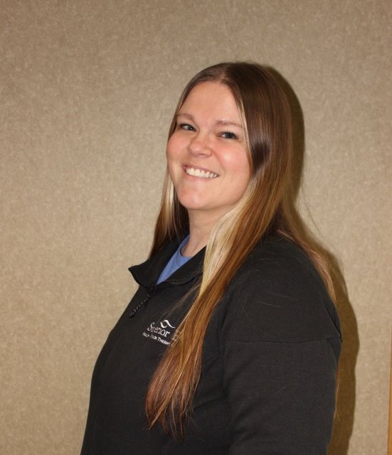 A young woman with long brown hair smiling at the camera, wearing a black jacket with a logo on it, standing against a plain beige wall.