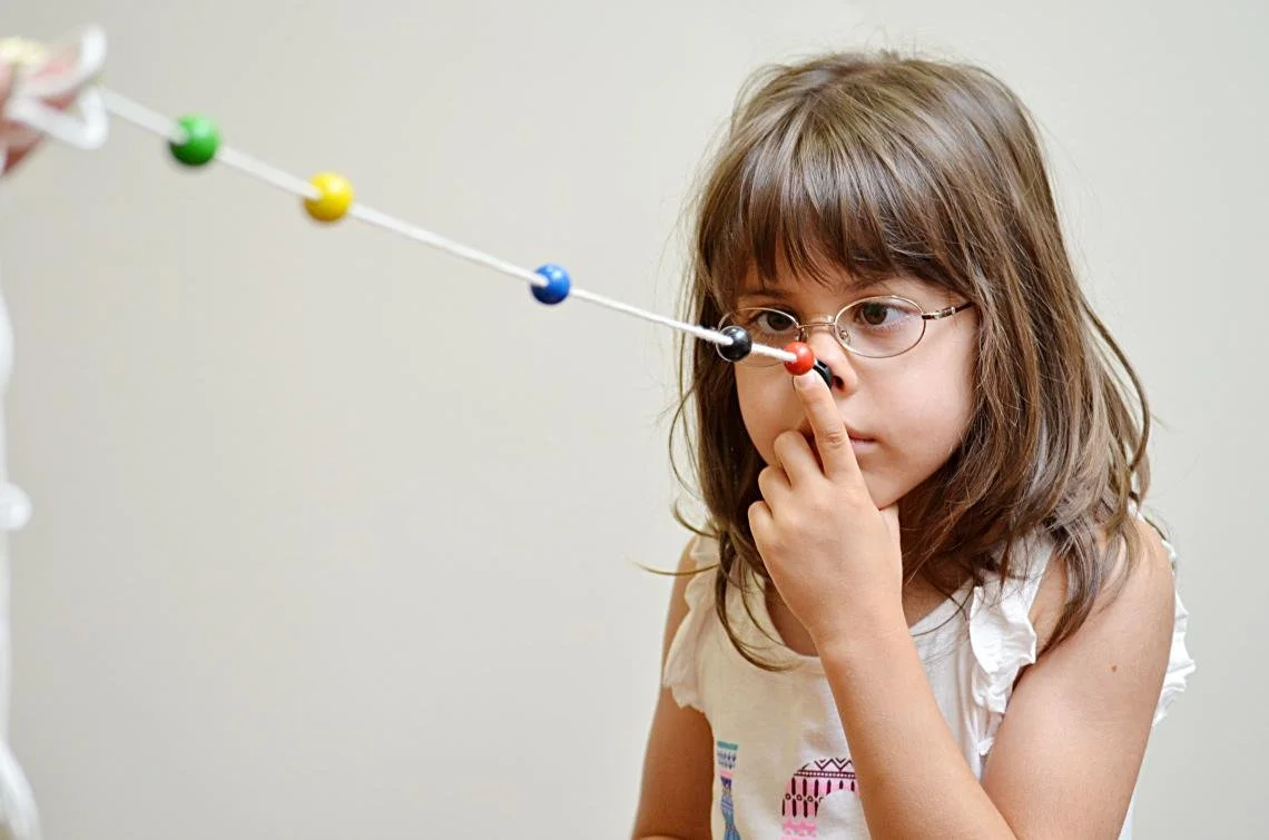 A young girl with glasses and brown hair pinches her nose while looking at a model of a molecule or atom structure with colorful balls on white sticks. She is doing a Vision Therapy exercise.