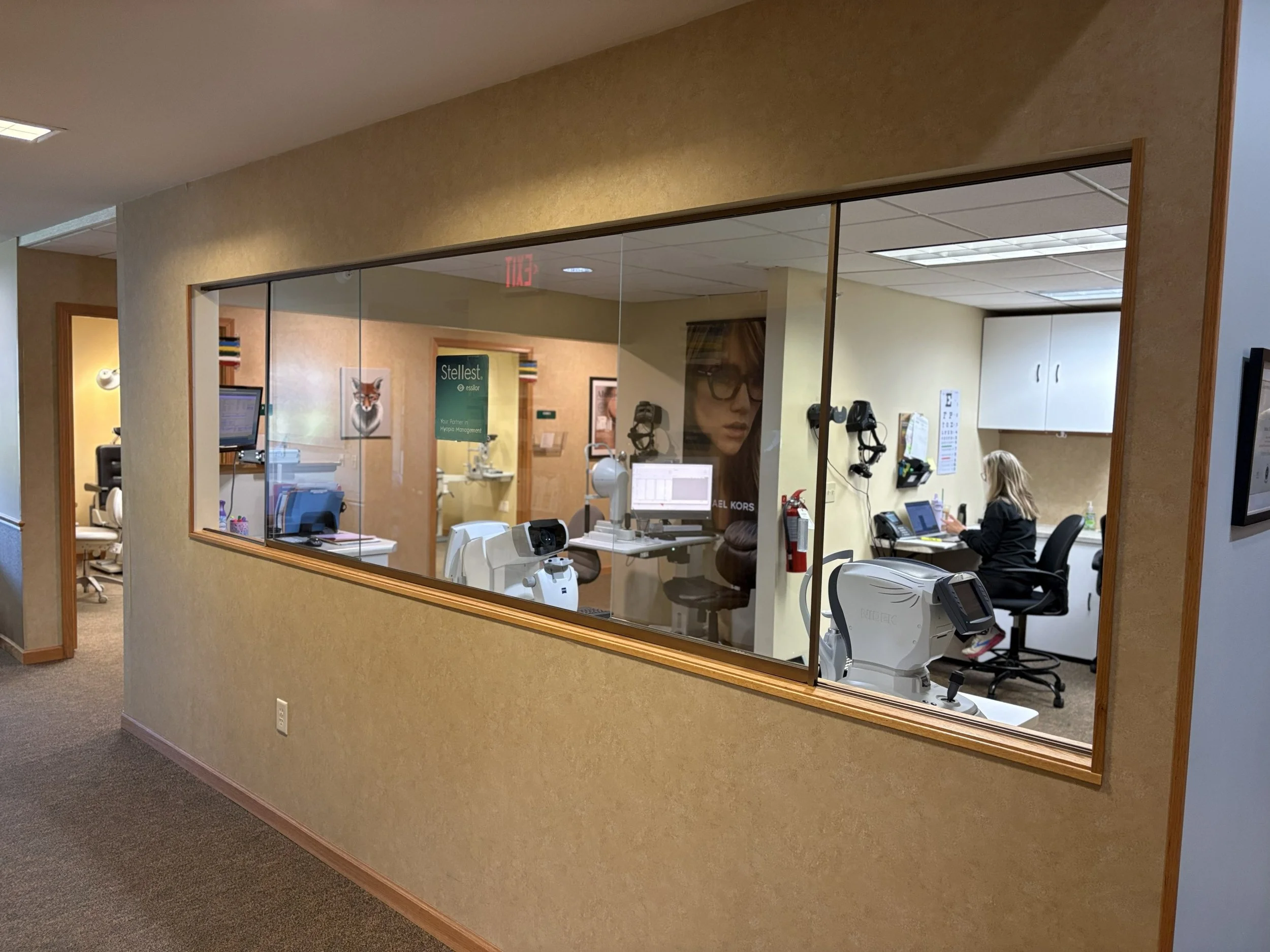 A view through a large glass window into a medical or dental office with a woman working at a desk, medical equipment, and a wall with books and posters.