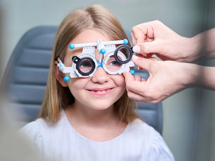 A young girl at an eye exam wearing a trial frame used for vision testing, with an eye doctor adjusting the device. She is doing a Vision Therapy exercise.