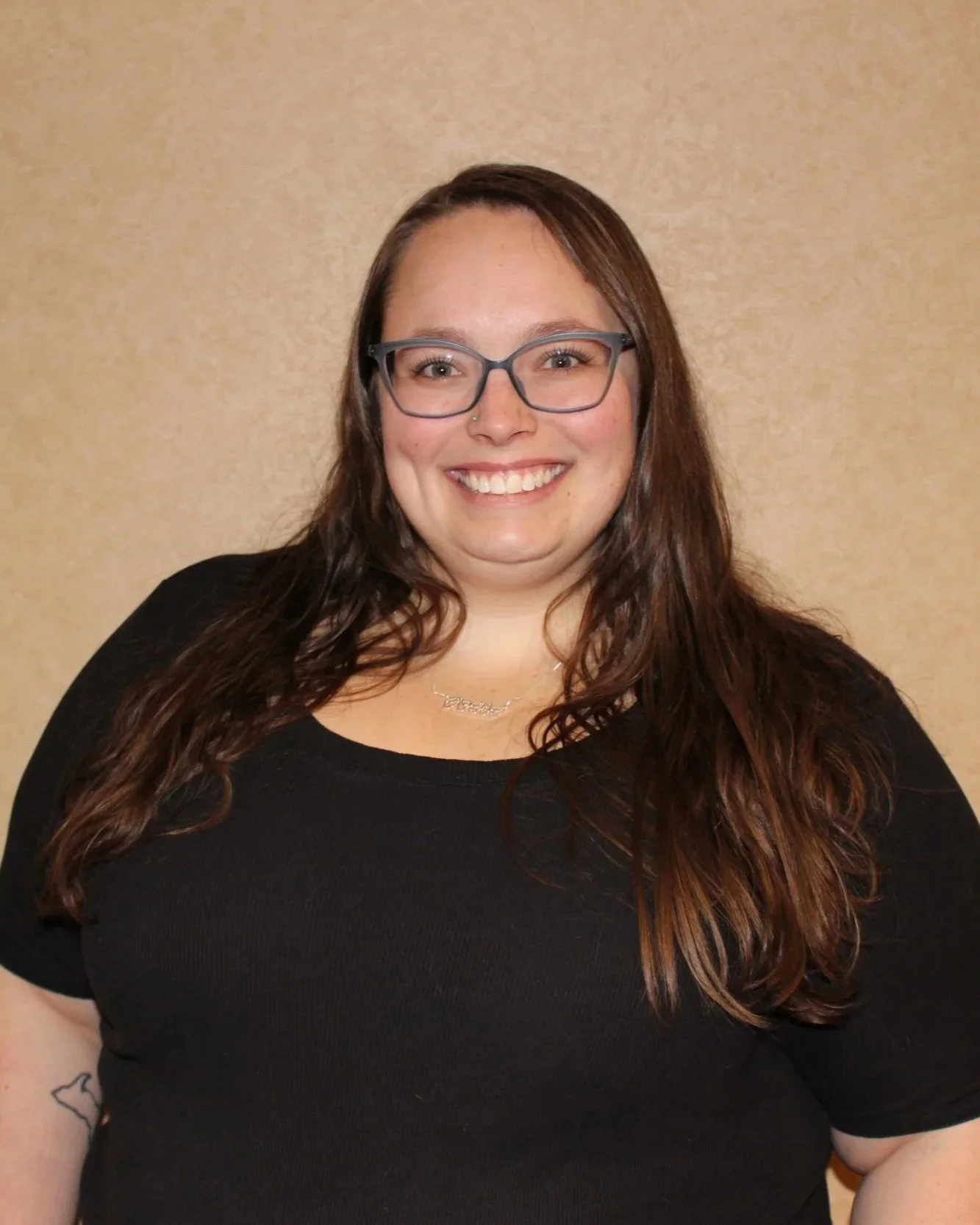 A woman with long wavy brown hair, wearing glasses, a black top, and a necklace, smiling at the camera, against a beige background.