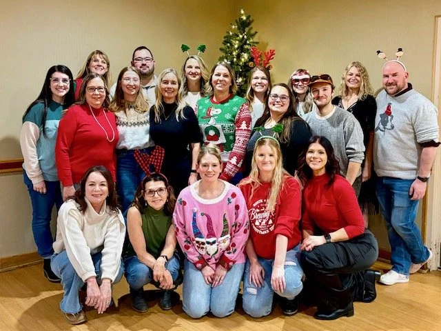 Group of 15 people posing for a photo during a holiday celebration, standing in front of a decorated Christmas tree in an indoor setting.