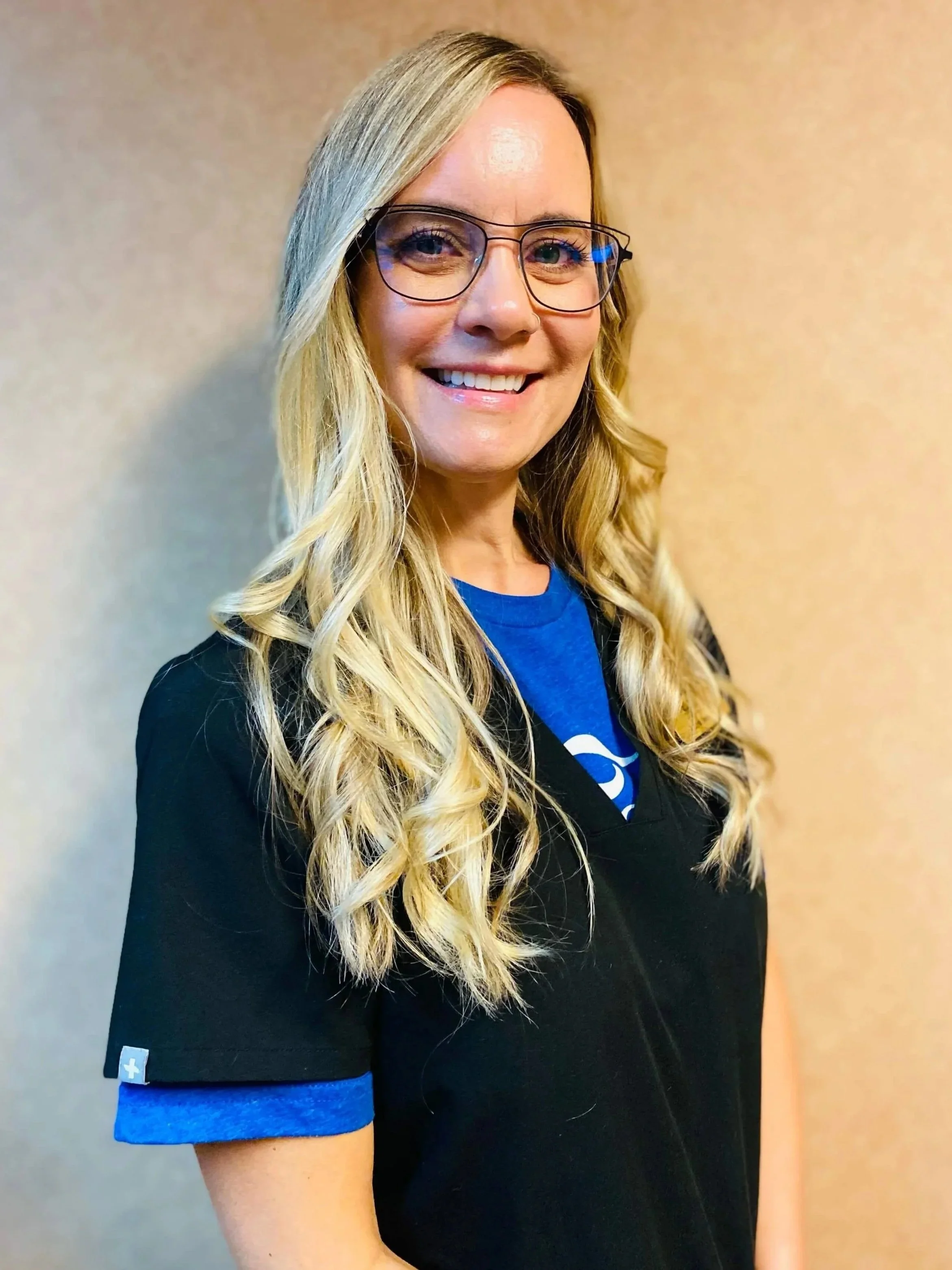 A woman with long blonde hair, glasses, and a big smile, wearing a black top and blue shirt, standing against a beige wall.
