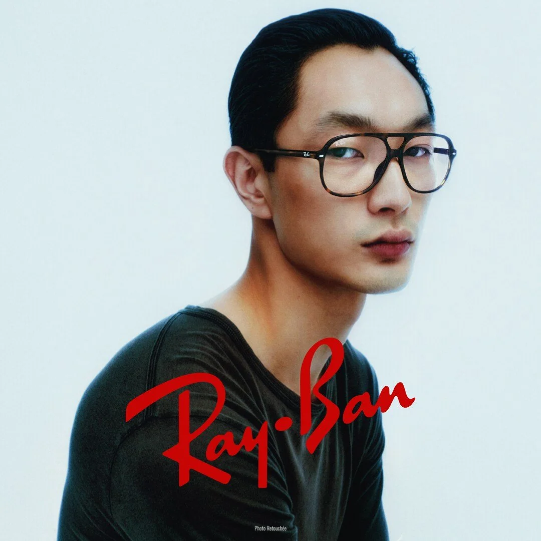 A young man with glasses and dark hair, wearing a black shirt, looking at the camera against a plain white background with "Ray-Ban" branding in red text.