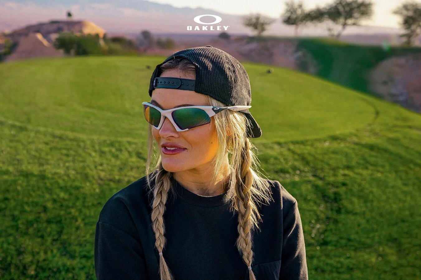A woman with blonde hair in braids wearing a black cap backwards, white Oakley sunglasses with green reflective lenses, and a black shirt, standing outdoors on a grassy hill at sunset.