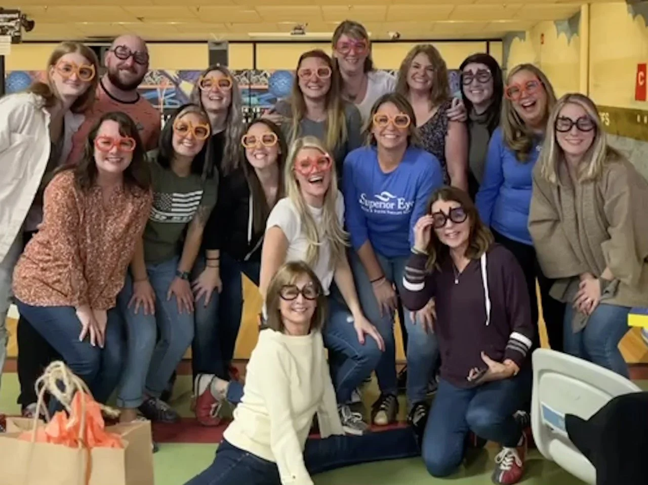 A group of 17 women and 1 man posing together indoors, all wearing colorful glasses, smiling, and appearing happy.