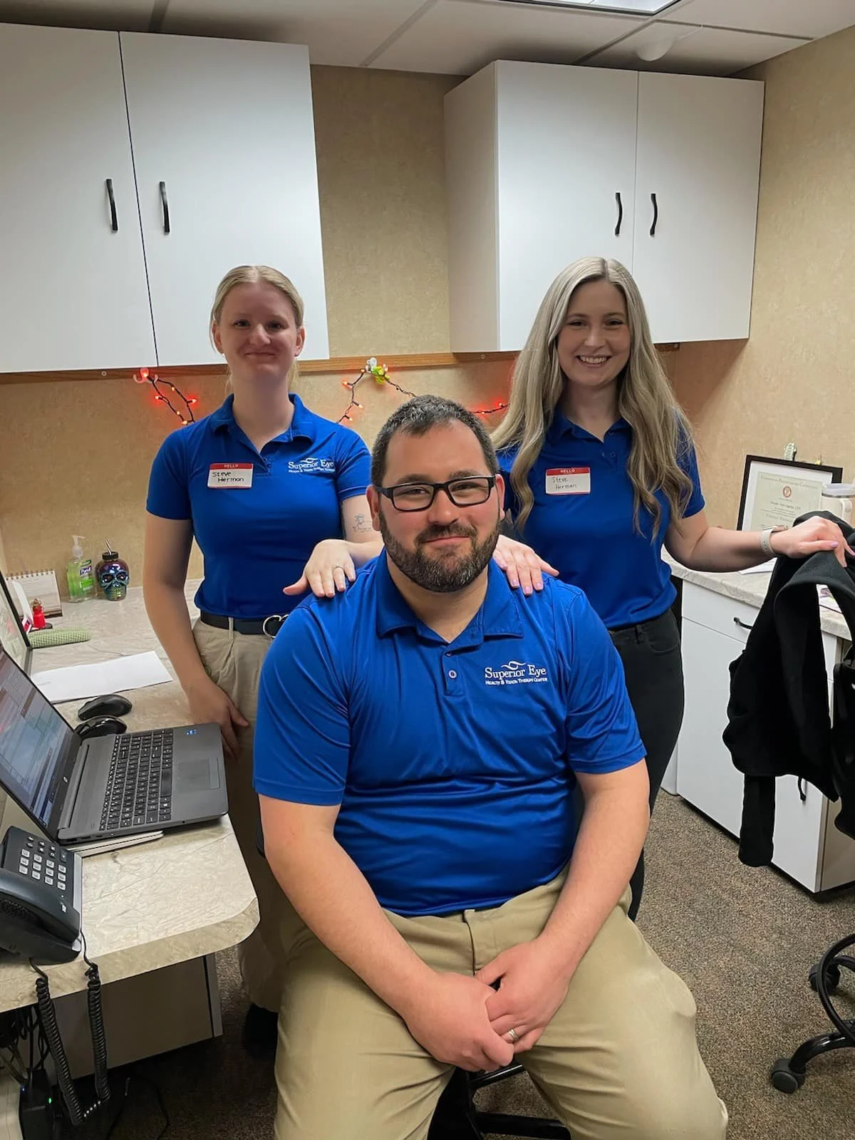 Three staff members in blue shirts with 'Superior Eye' logos, two women standing behind a seated man, all smiling, in an office with cabinets, desk, and holiday lights.