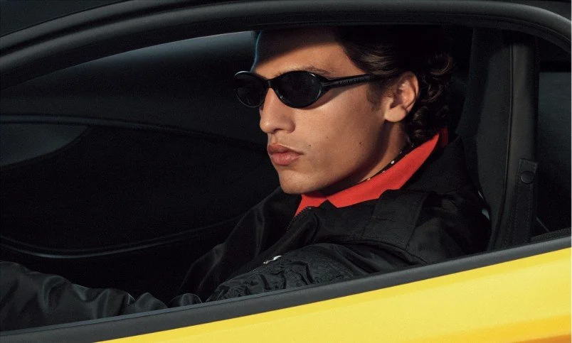 A man with curly hair wearing black sunglasses and a black jacket with red accents sitting in a yellow car.