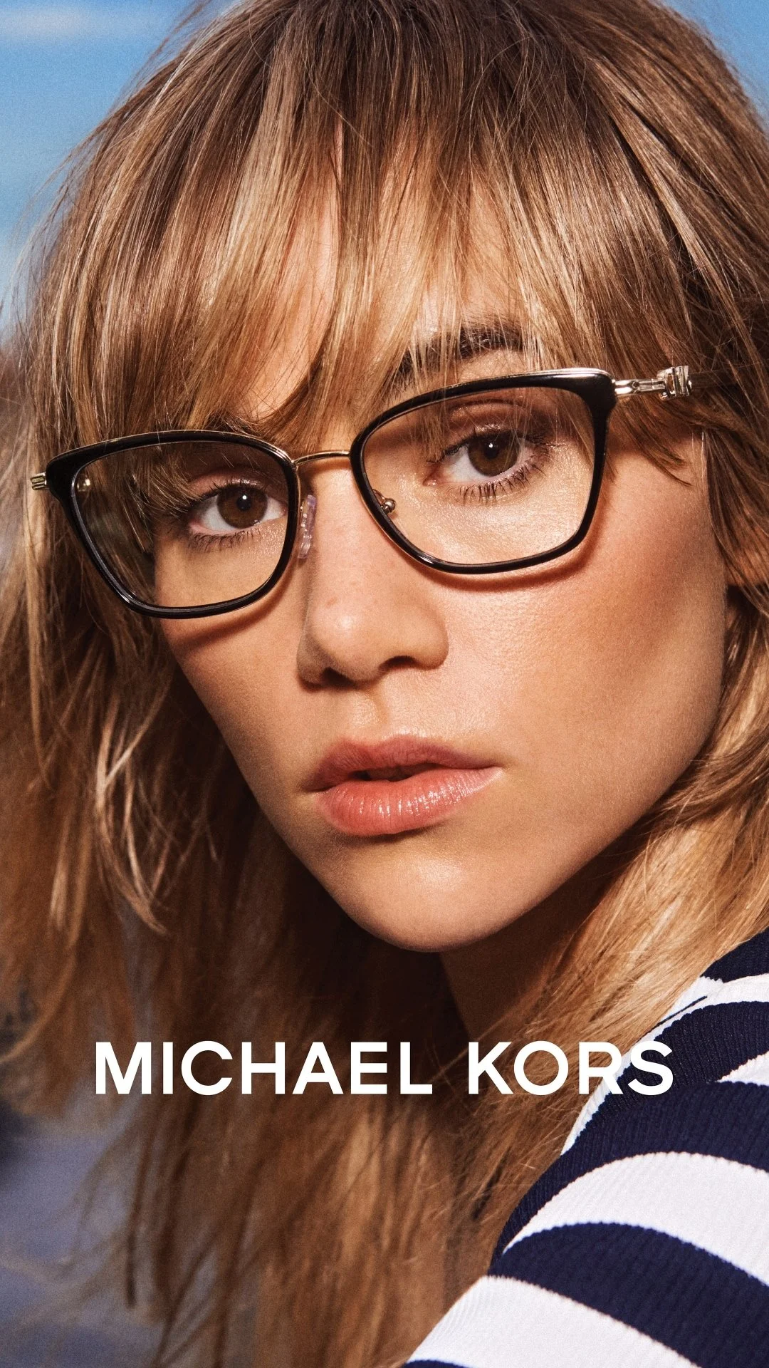 Close-up of a woman with light brown hair, wearing black-rimmed glasses, and a striped shirt, with the text 'MICHAEL KORS' at the bottom.