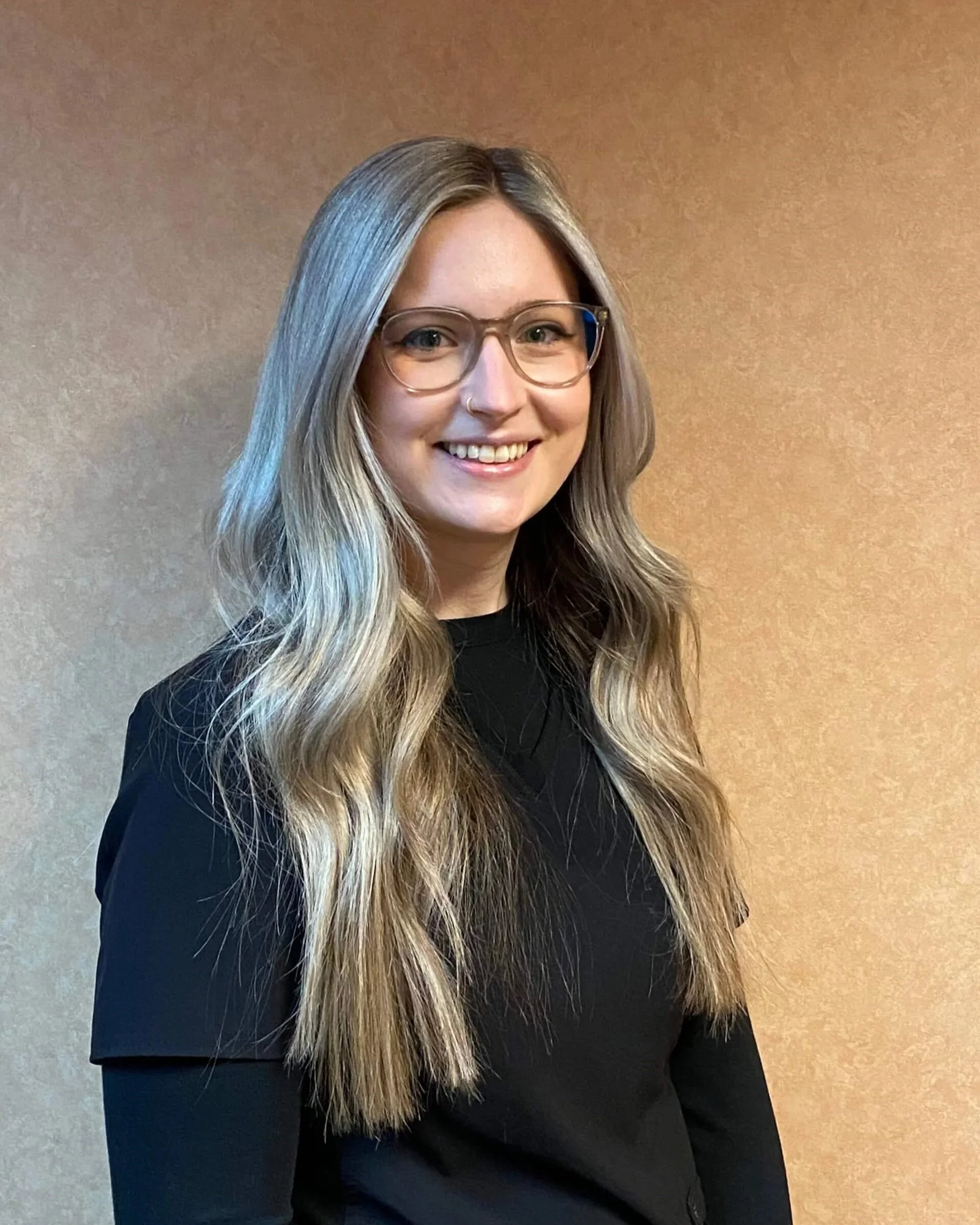 Portrait of a woman with long, wavy blonde hair wearing glasses and a black top, smiling and standing against a plain beige background.