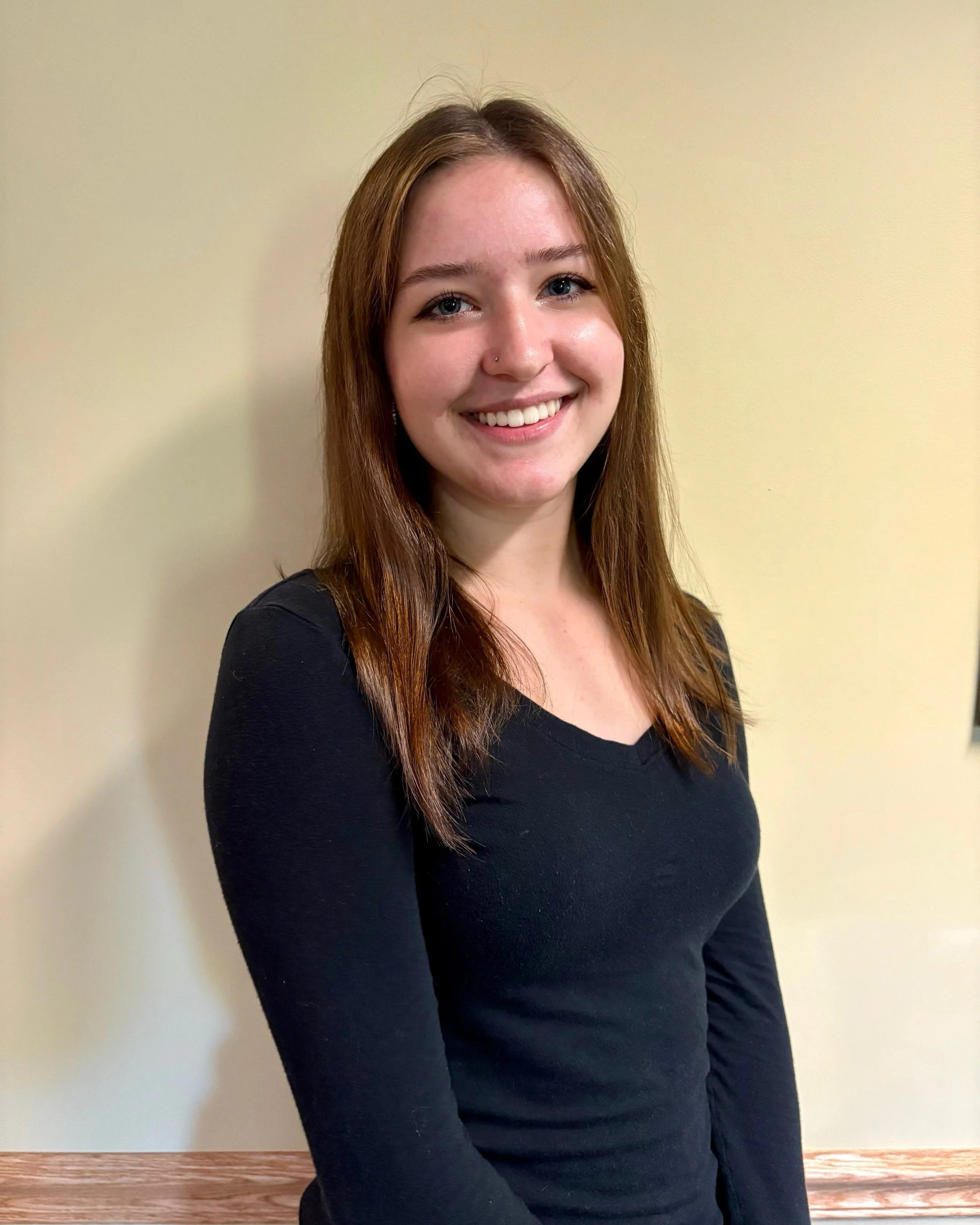 A young woman with long, reddish-brown hair and a nose piercing smiling, wearing a black long-sleeve top, standing against a beige wall and a wooden floor.