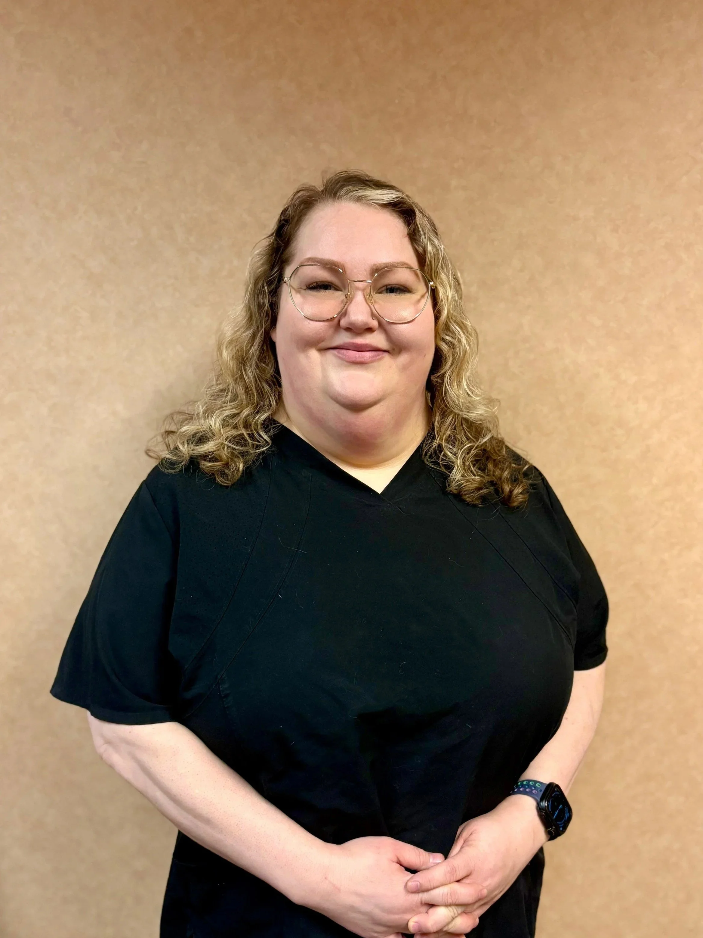 A woman with curly blonde hair wearing glasses and a black top, standing with hands clasped in front of her against a plain beige wall.