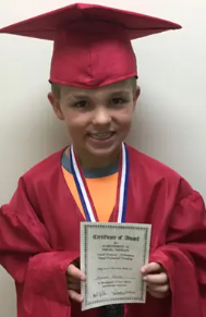 A young child in a red graduation cap and gown holding a diploma, wearing a medal around their neck, smiling. He is a Vision Therapy graduate from Superior Eye Health.