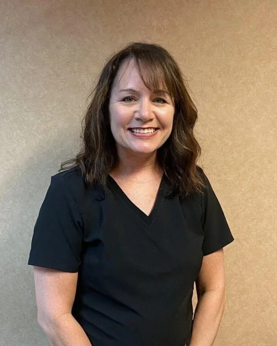 A woman with shoulder-length brown hair smiling, wearing a black V-neck shirt, standing against a beige wall.